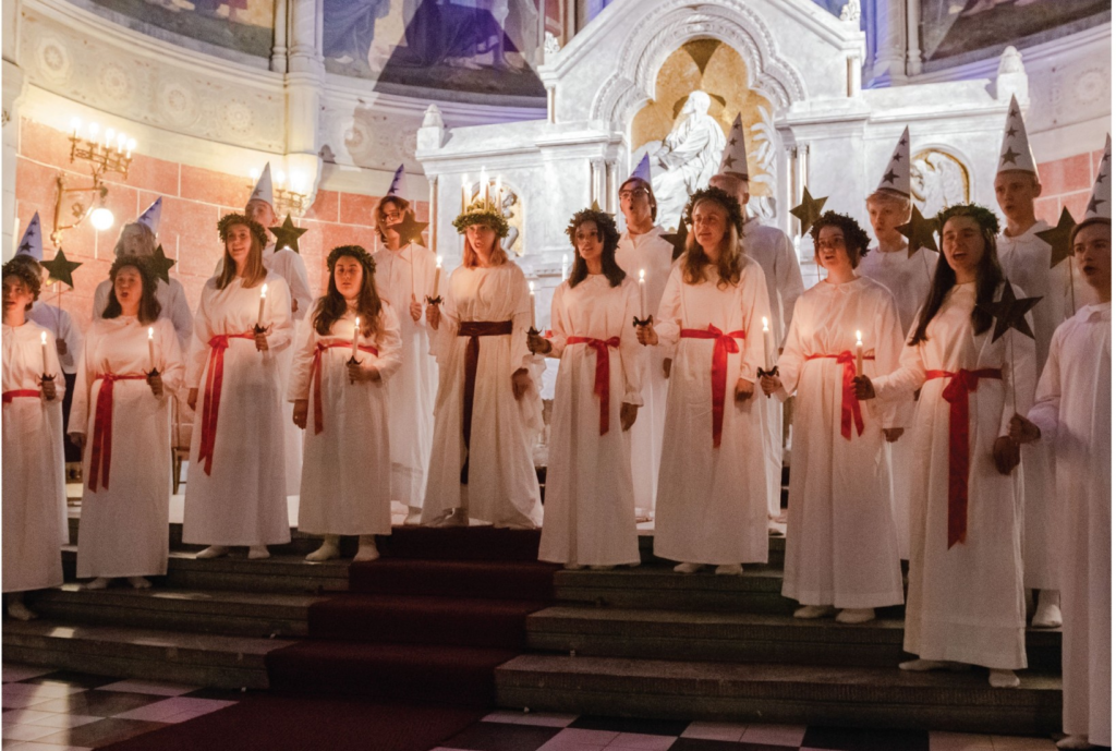 zdjęcie chóru. dziewczęta i chłopcy ubrani w białe tuniki, przepasani czerwonymi szarfami. W ręce trzymają świece. Stoją na schodach we wnętrzu kościoła